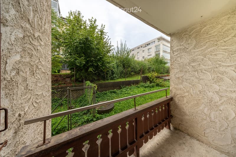 Balcony with wooden railing and metal handrail, overlooking a green, overgrown area with trees and a building in the background.