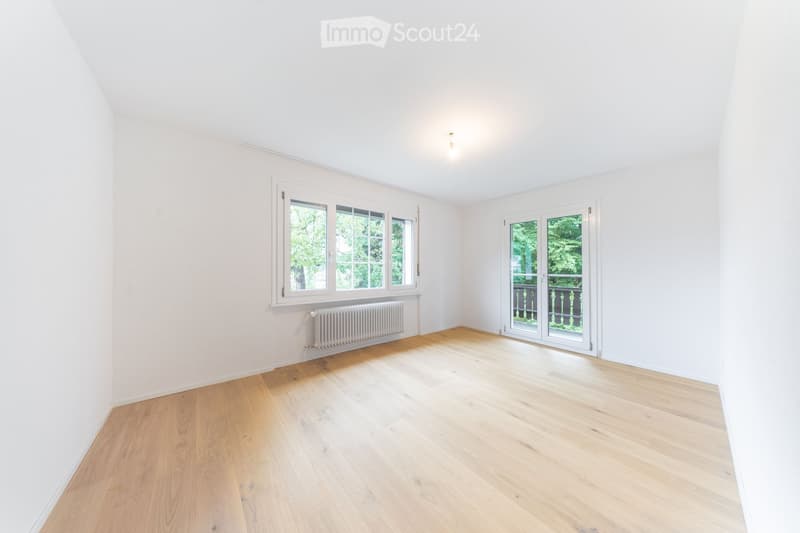 Empty room with light wooden flooring, white walls, large windows, and a radiator. The room has a sliding glass door leading to a balcony or outdoor area.