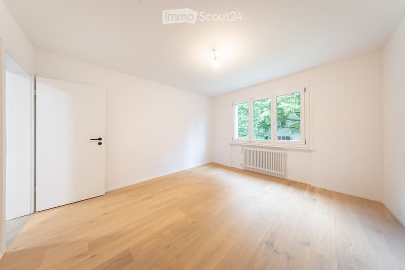 Empty room with light wooden flooring, white walls, large windows, and a radiator. No furniture or equipment present.