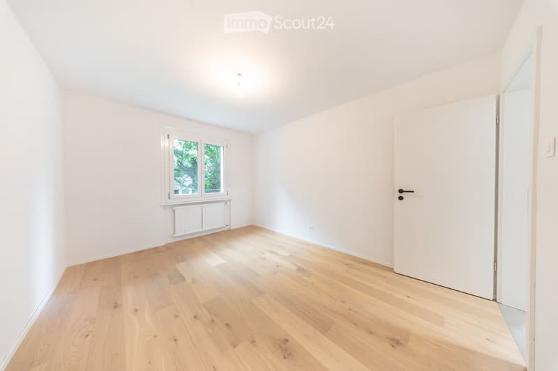 Empty room with light wooden flooring, white walls, a window with white frame, radiator, and a white door.