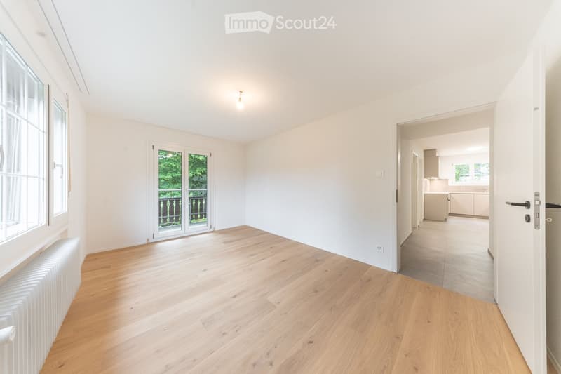 Empty room with light wooden flooring, white walls, large windows, and a radiator. A doorway leads to a kitchen area with white cabinets and countertops.