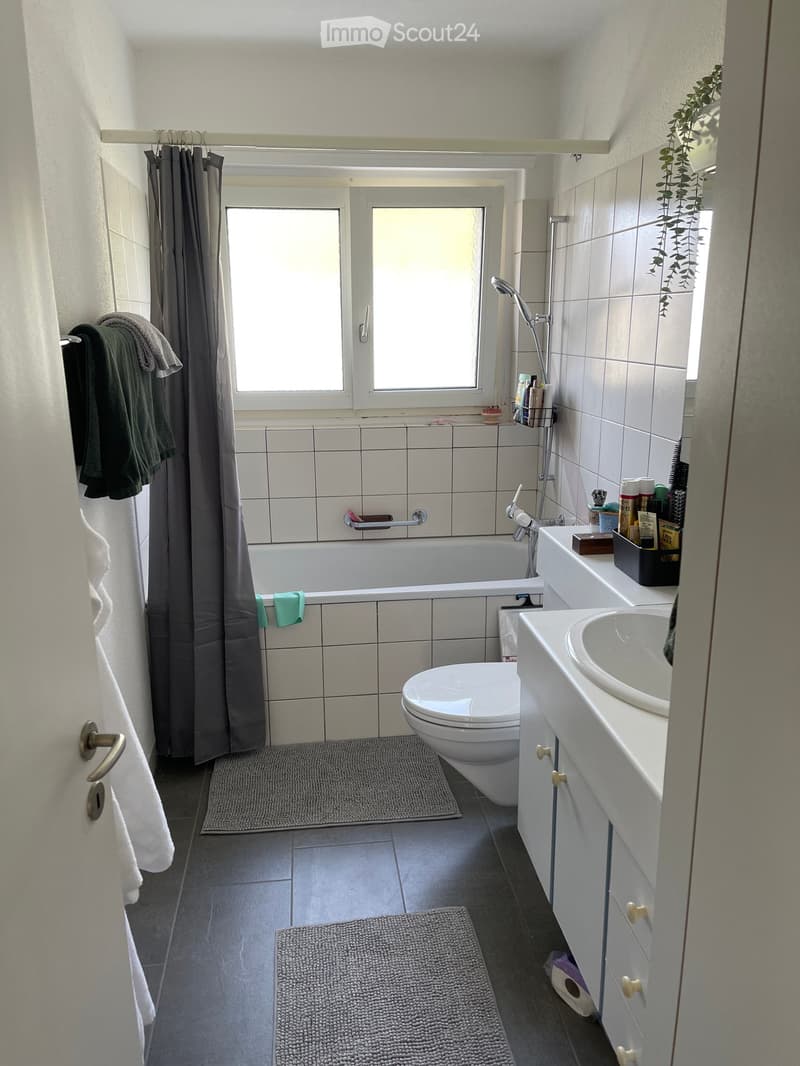 Modern bathroom with a bathtub, toilet, sink, and bathroom cabinet. Features tiled walls, a window with a shower curtain, and towels on a rack.