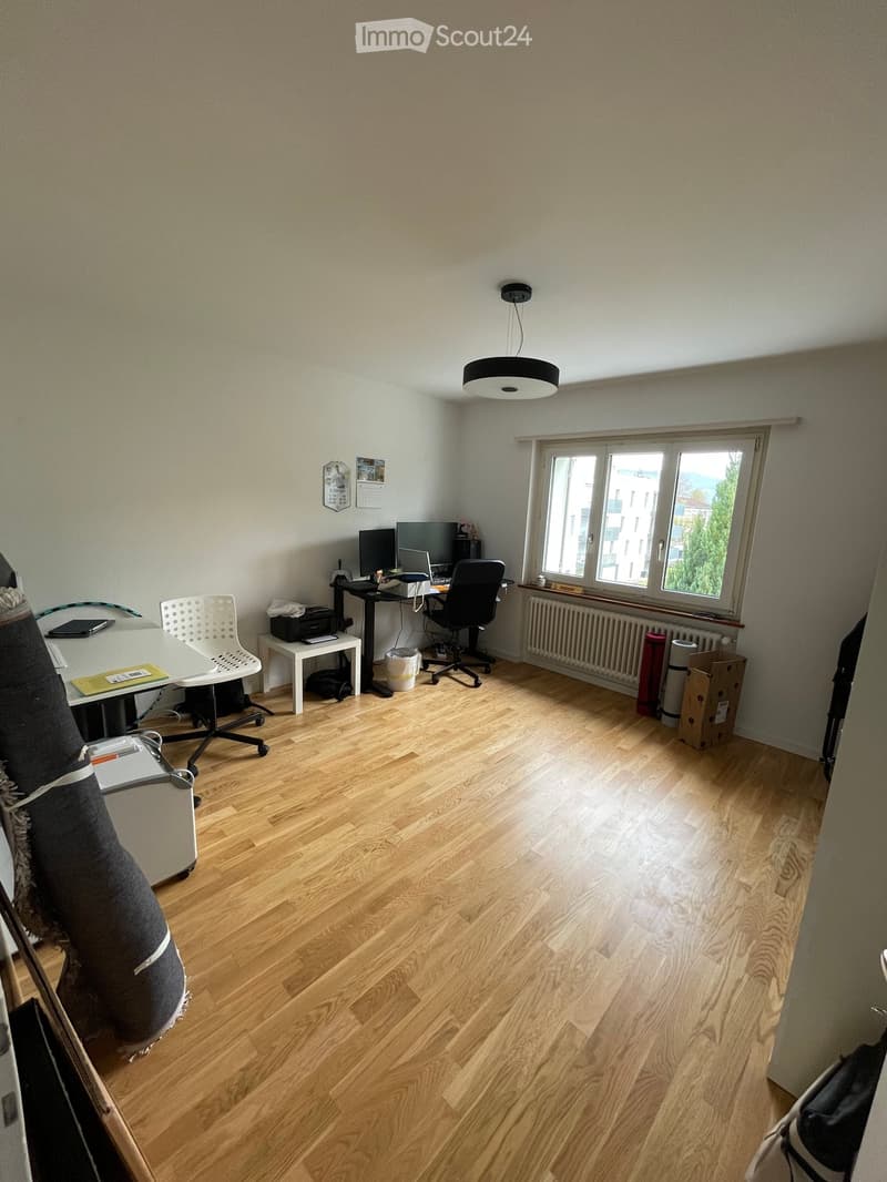 A furnished room with wooden flooring, a large window, a desk with a laptop, a chair, and a printer. There is also a radiator under the window and a small table with a chair.