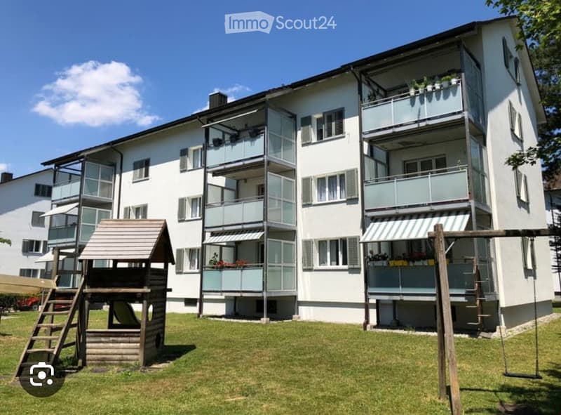 A modern multi-story residential building with white exterior, multiple balconies, and a playground area in front.