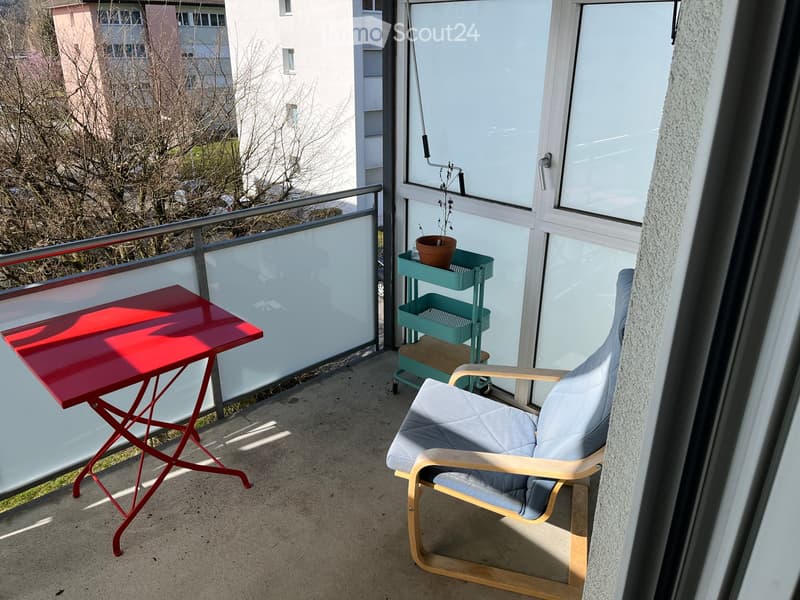 Balcony with a red folding table, a grey upholstered chair, a small green cart with a potted plant, and a glass railing overlooking a residential area.
