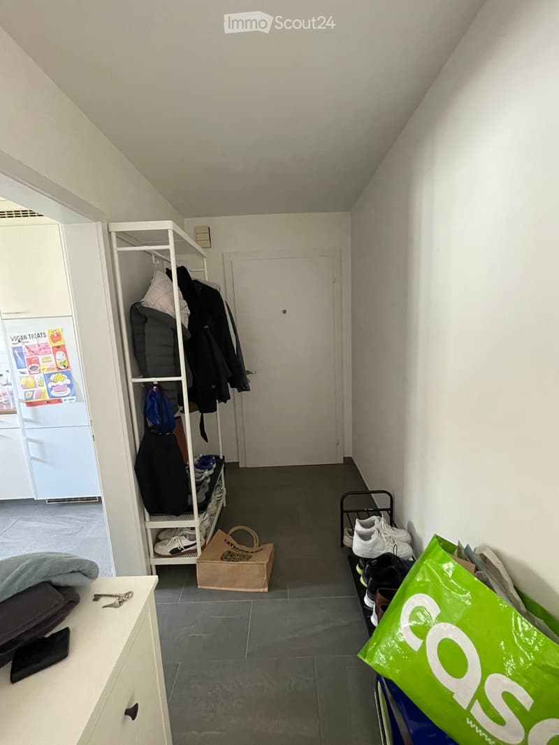 A narrow hallway with white walls and a gray tiled floor. A white coat rack holds jackets and shoes, a small table with folded blankets and keys, and a green shopping bag is visible.