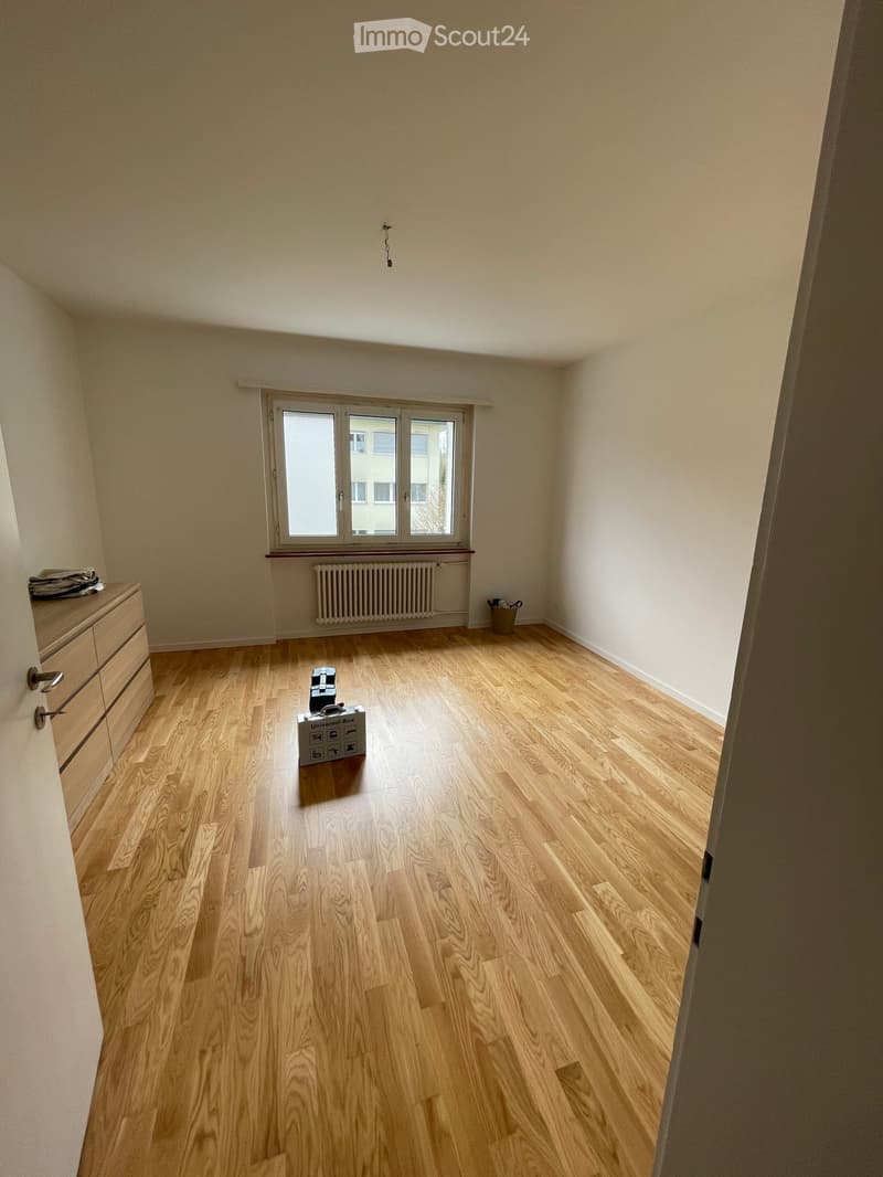 Empty room with wooden flooring, white walls, and a window with a radiator below it. A small box and a few items are placed on the floor.