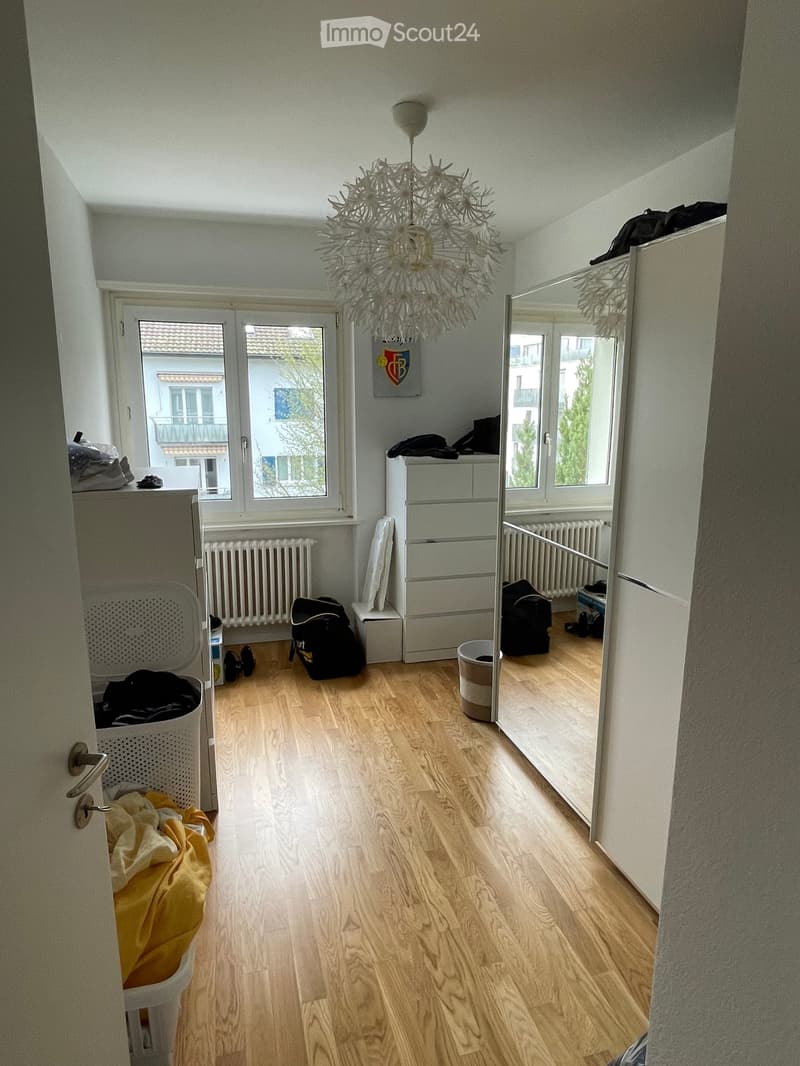 Bright hallway with wooden flooring, large windows, and a modern chandelier. Visible items include a laundry basket, a mirror, and some bags.