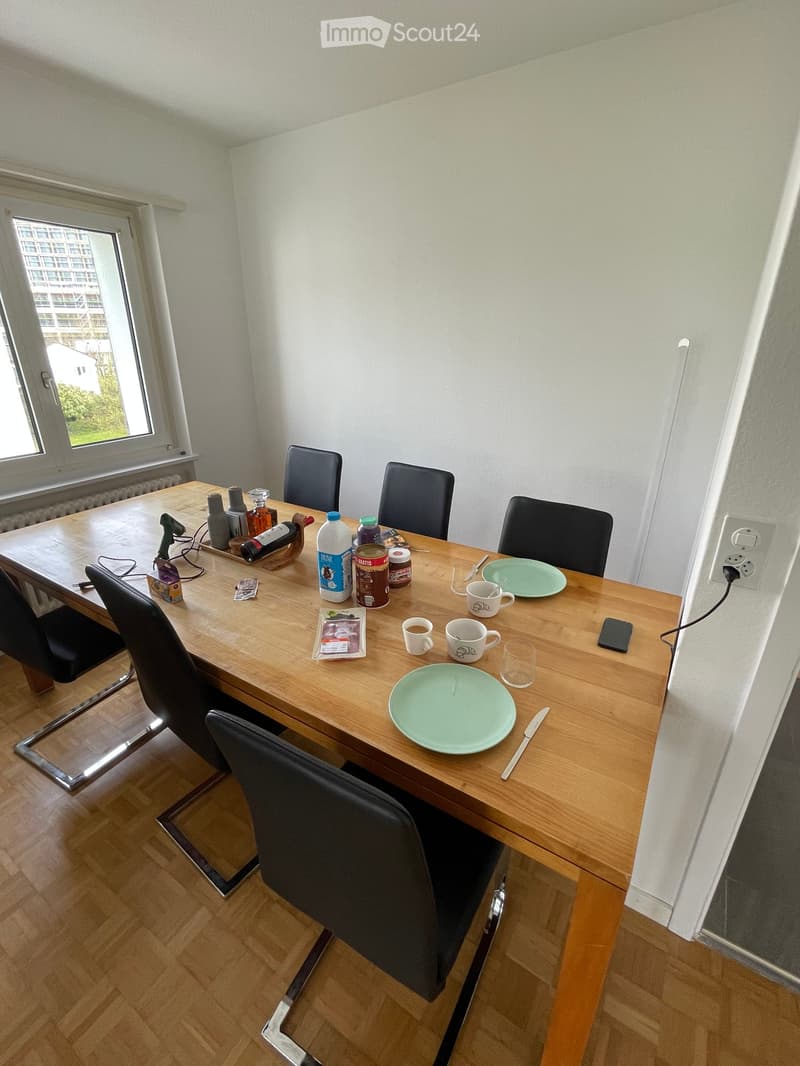 A wooden dining table with four black chairs, set with plates, cups, and various items including a bottle and a cellphone. The room has light-colored walls and wooden flooring.