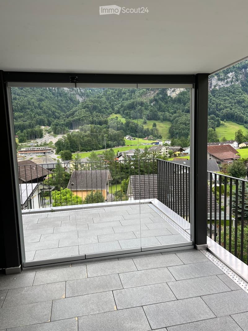 Modern balcony with large glass windows offering a scenic view of a green valley and houses. The floor is tiled, and the balcony is enclosed with a sleek black metal railing.