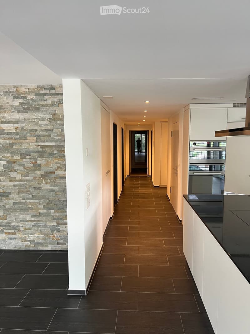 Modern hallway with a stone accent wall, wooden flooring, white cabinetry, and recessed lighting. The hallway leads to a kitchen area with stainless steel appliances and a black countertop.