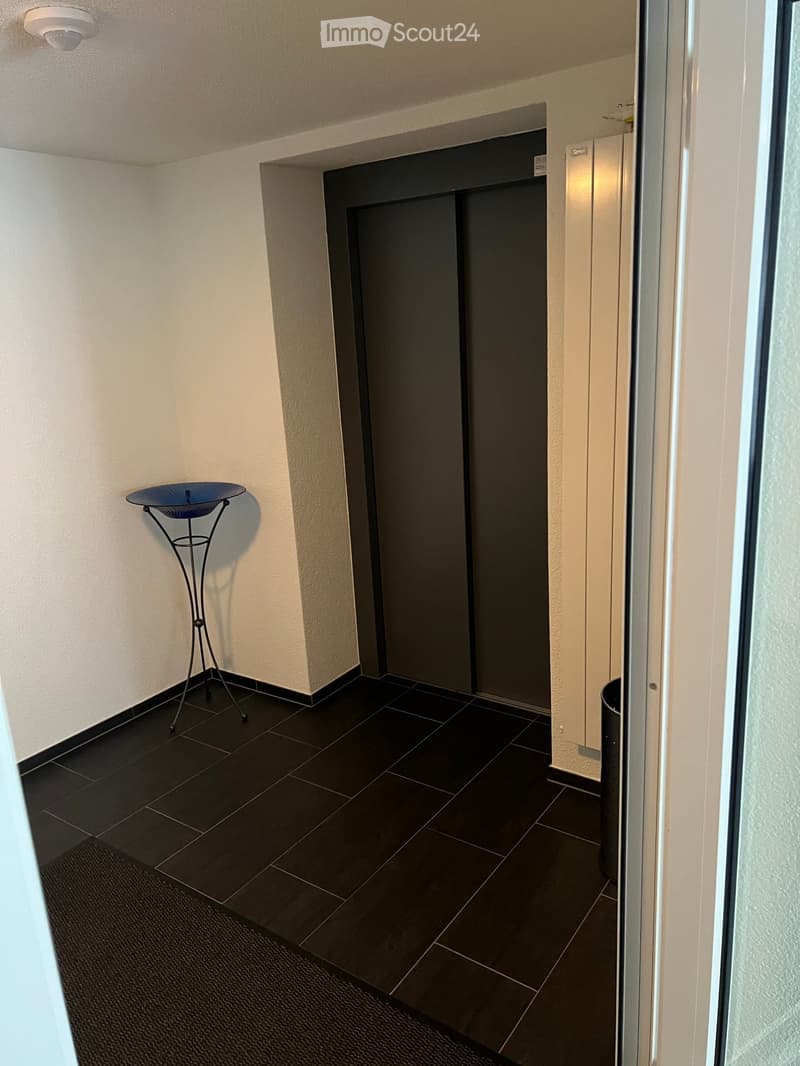 Hallway with dark tiled floor, white walls, elevator, small decorative table, and trash bin.