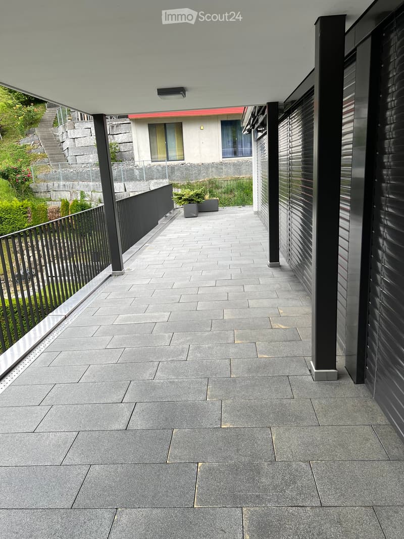 Modern balcony with grey tiled flooring, black metal railing, and a view of a landscaped garden area with steps and plants.