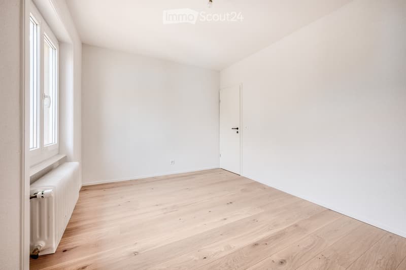 Empty room with light wooden flooring, white walls, a white radiator, and a closed door.