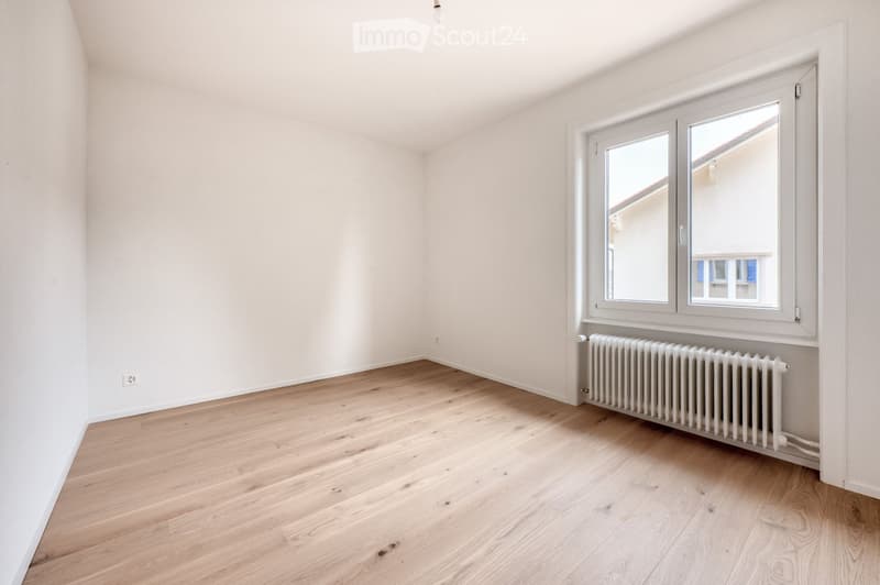 Empty room with light wooden flooring, white walls, and a large window with white frames. A radiator is visible beneath the window.