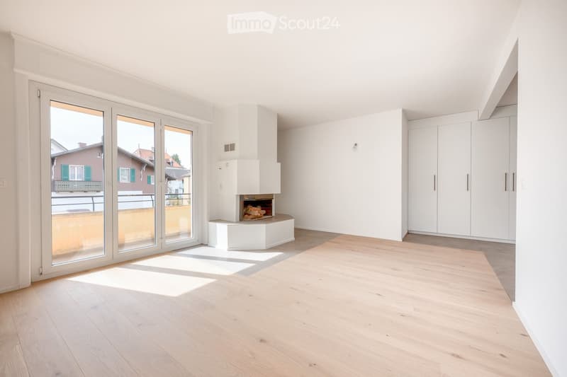 Bright living room with light wooden flooring, white walls, built-in white cabinets, and a modern fireplace. Large glass sliding doors lead to a balcony with a view of neighboring houses.