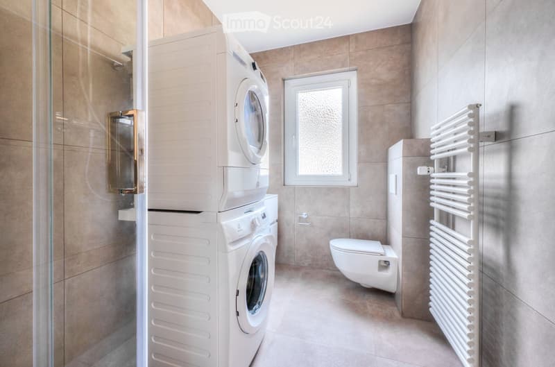 Modern bathroom with beige tiles, a stacked washing machine and dryer, a toilet, a white radiator, and a glass shower enclosure.