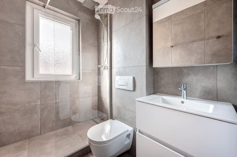 Modern bathroom with a toilet, sink, and shower area. Features include a glass-enclosed shower, a modern faucet, and a sleek white cabinet.