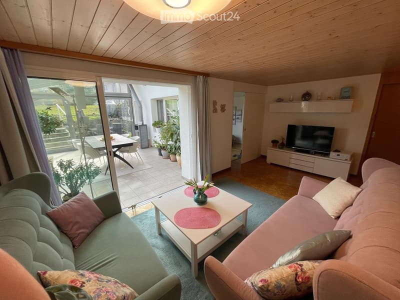 Bright living room with wooden ceiling, large windows, and a view to the garden. Features a green sofa, pink sofa, coffee table with a vase, and a TV on a white stand.