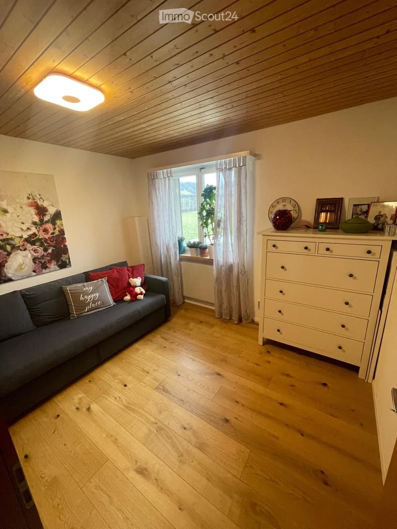 Bright living room with wooden flooring, a grey sofa with red cushions, a white drawer unit, and a large window with curtains.