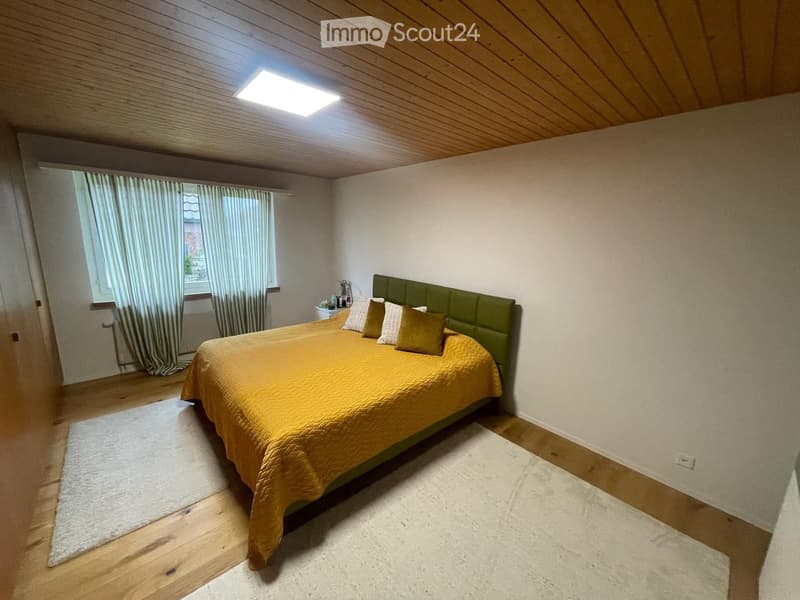 Bedroom with wooden flooring, slanted wooden ceiling, large window with curtains, and a neatly made bed with pillows and a nightstand.