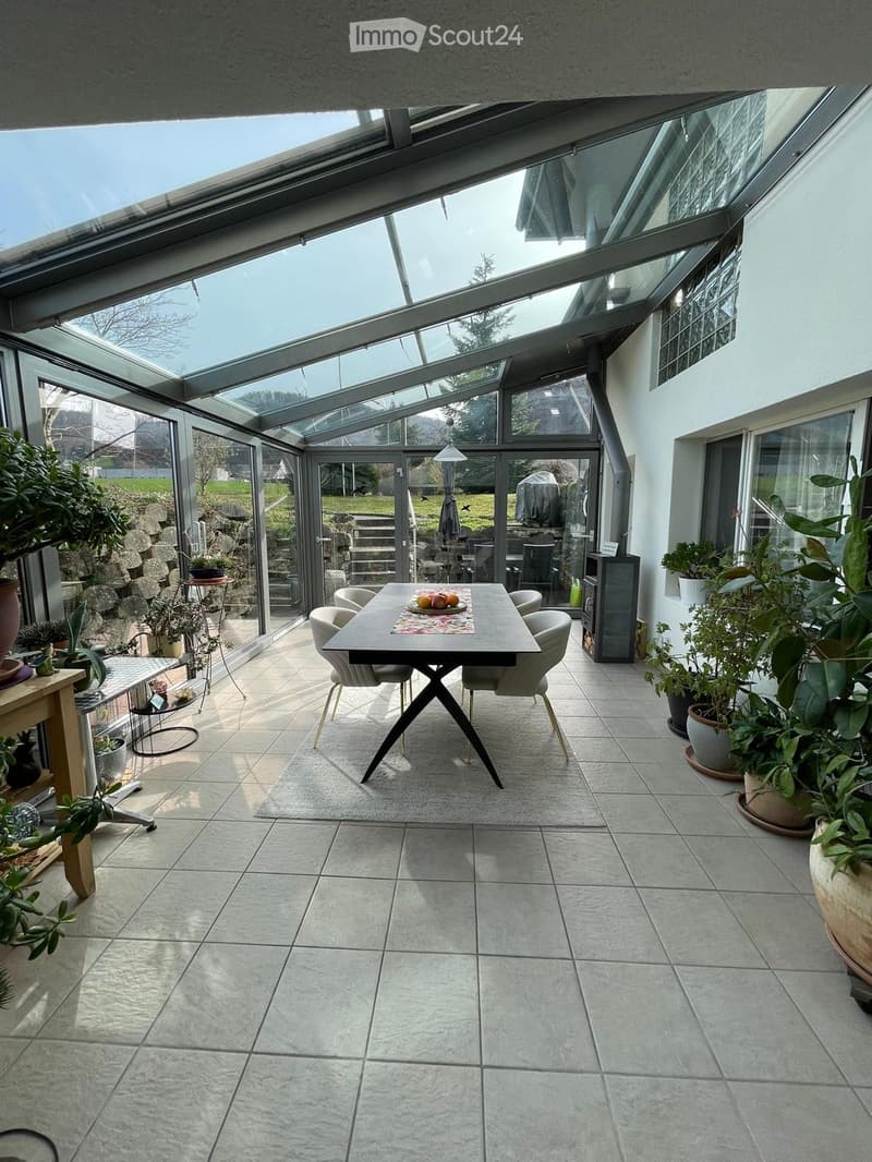 Bright, airy dining area with large glass walls and roof, featuring a modern dining table, chairs, side table, and several potted plants.