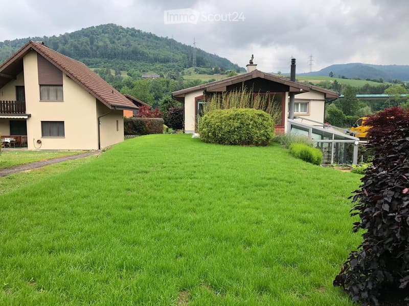 Two-story house with a red-tiled roof, surrounded by a well-maintained green lawn and bushes. The house features a balcony and a small patio area with outdoor furniture. The background shows a hilly landscape with trees and distant mountains.