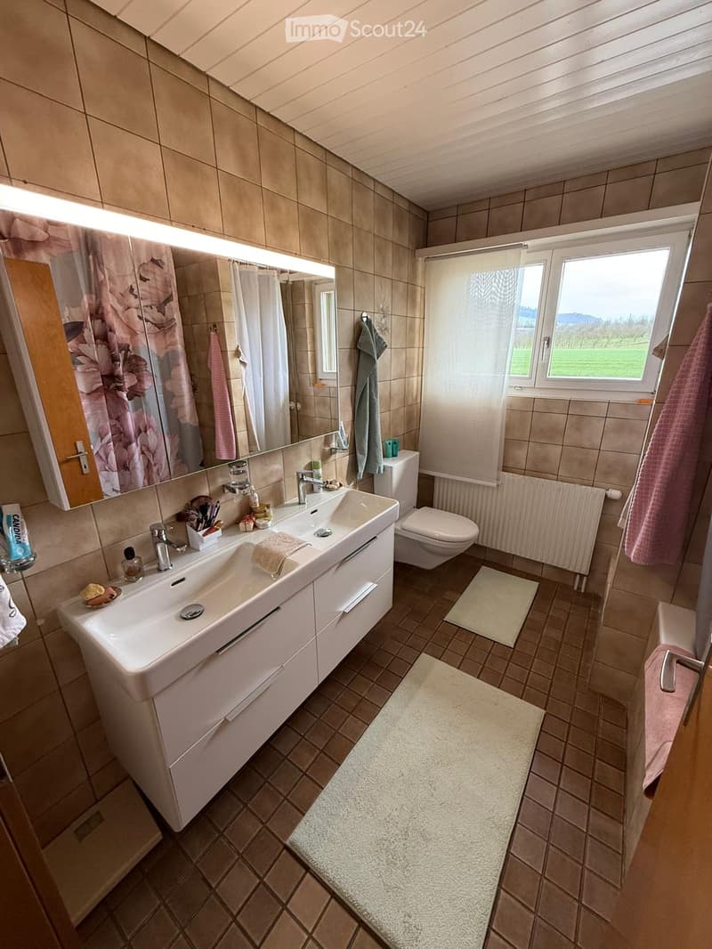Modern bathroom with beige tiles, double sink, large mirror, toilet, and towels. Features a window with a view of greenery.