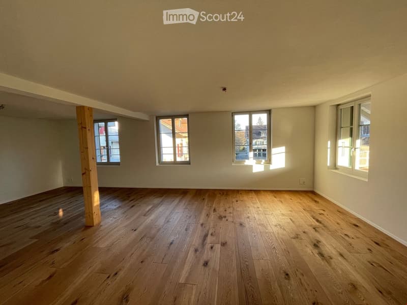 Empty room with wooden flooring and multiple windows, featuring light-colored walls and a wooden support beam.