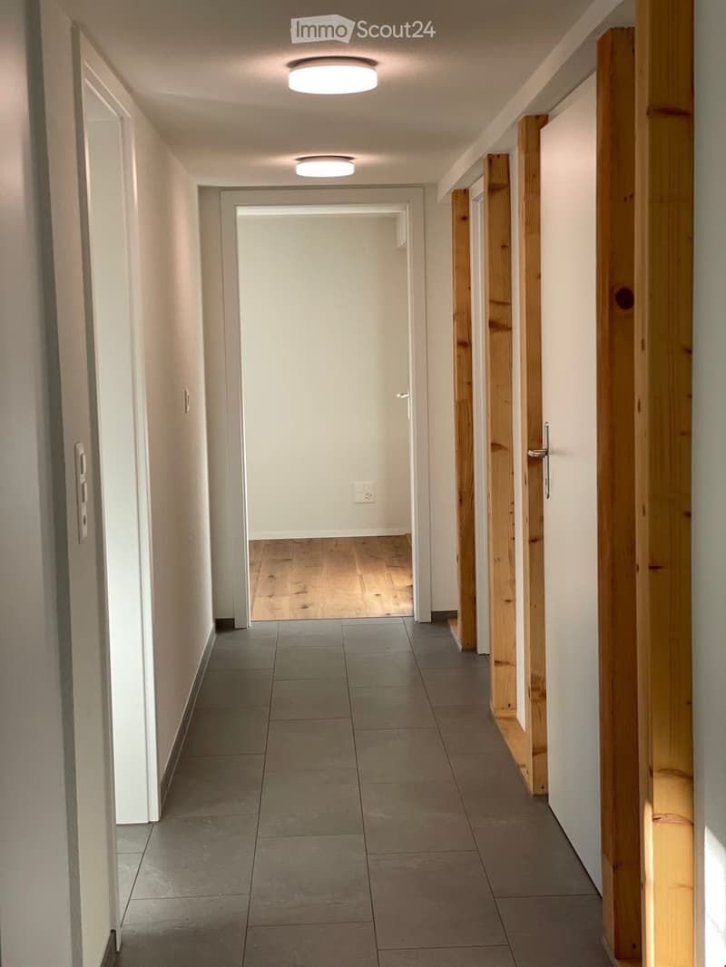 Clean, modern hallway with tiled floor, white walls, and recessed lighting. Wooden door frames and a visible room with wooden flooring at the end.