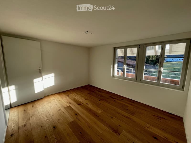 Empty room with wooden flooring, white walls, and large windows with grey frames. The room has a white door on the left side and natural light coming through the windows.
