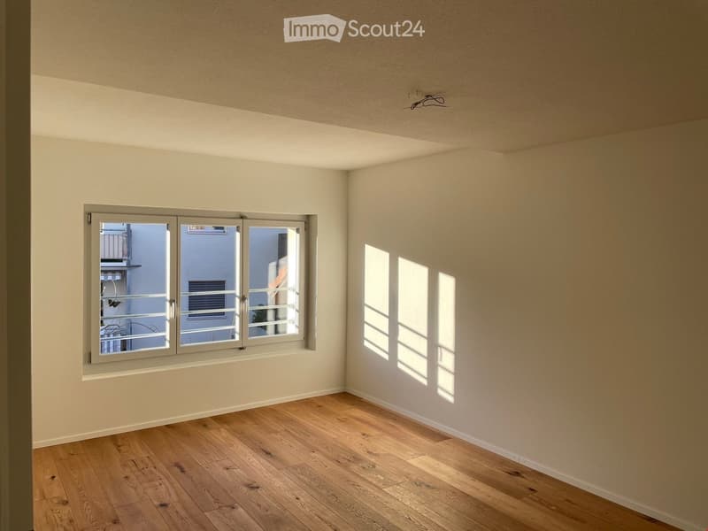 Empty room with light wooden flooring and white walls, featuring a large window with a view of a balcony and neighboring buildings.