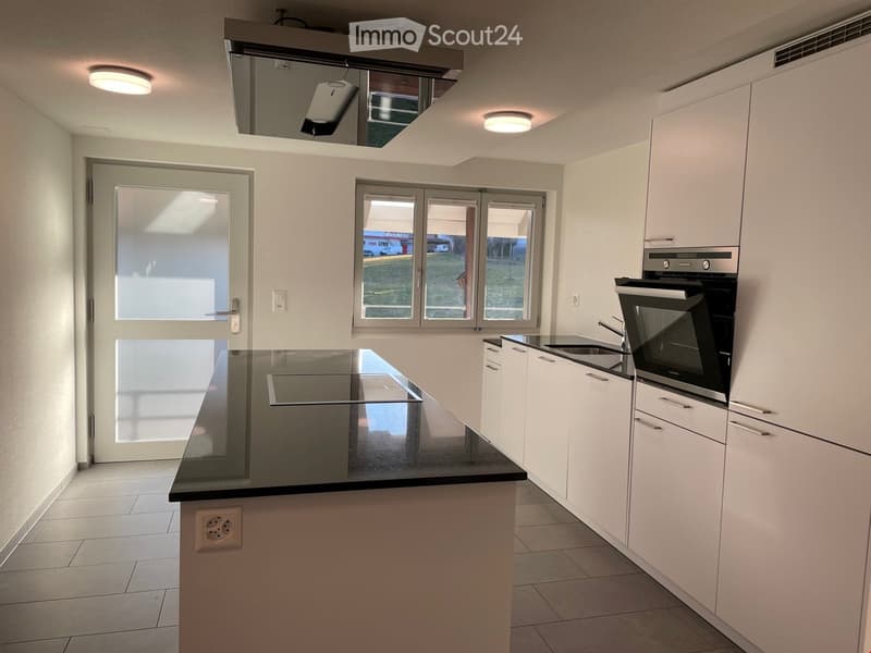Modern kitchen with white cabinets, black countertop, built-in oven, and island. Features large windows and a door leading outside.
