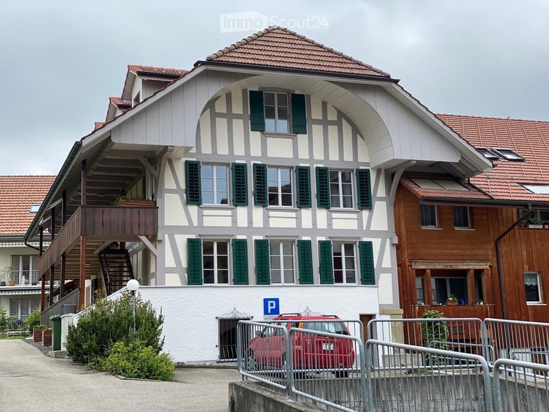 Two-story residential building with white and grey facade, green shutters, and red-tiled roof. Features a balcony on the upper floor and a parking area with a red car.