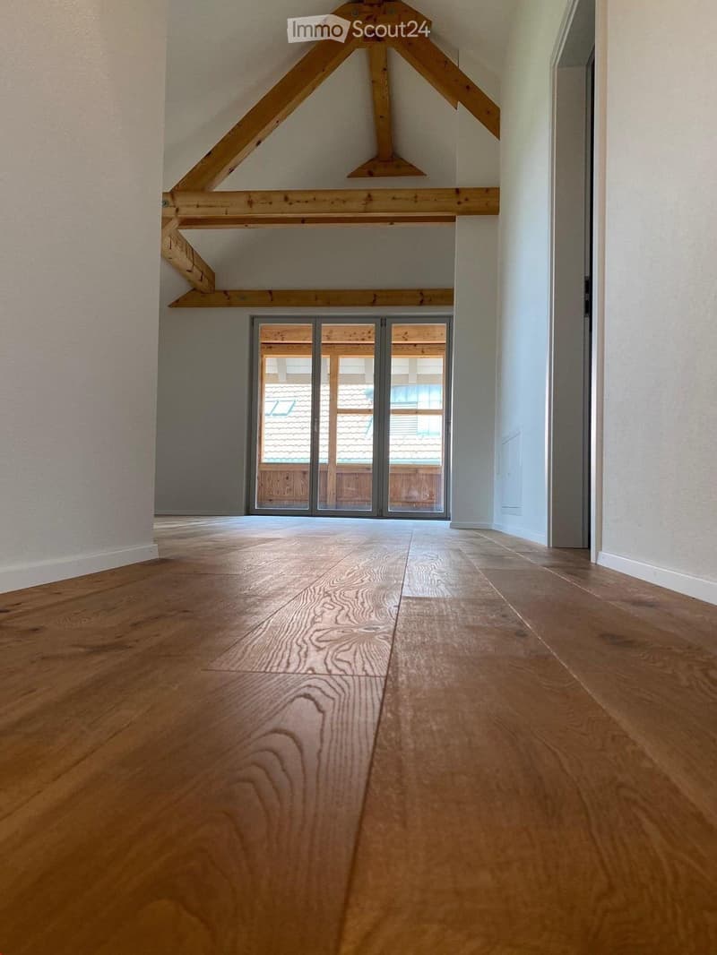 Empty hallway with wooden flooring, white walls, and a large sliding glass door leading to an outdoor area with visible wooden beams and a roof.