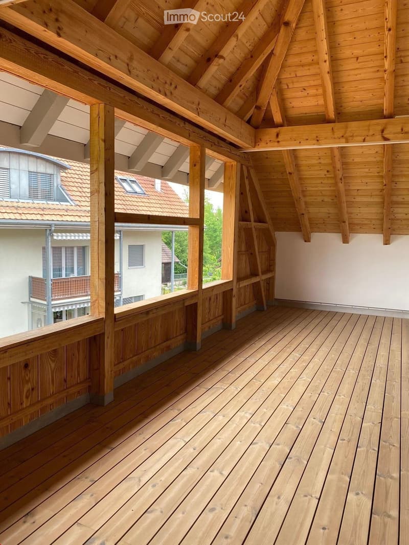 Wooden balcony with exposed wooden beams and railings, overlooking a residential area with neighboring houses and greenery.