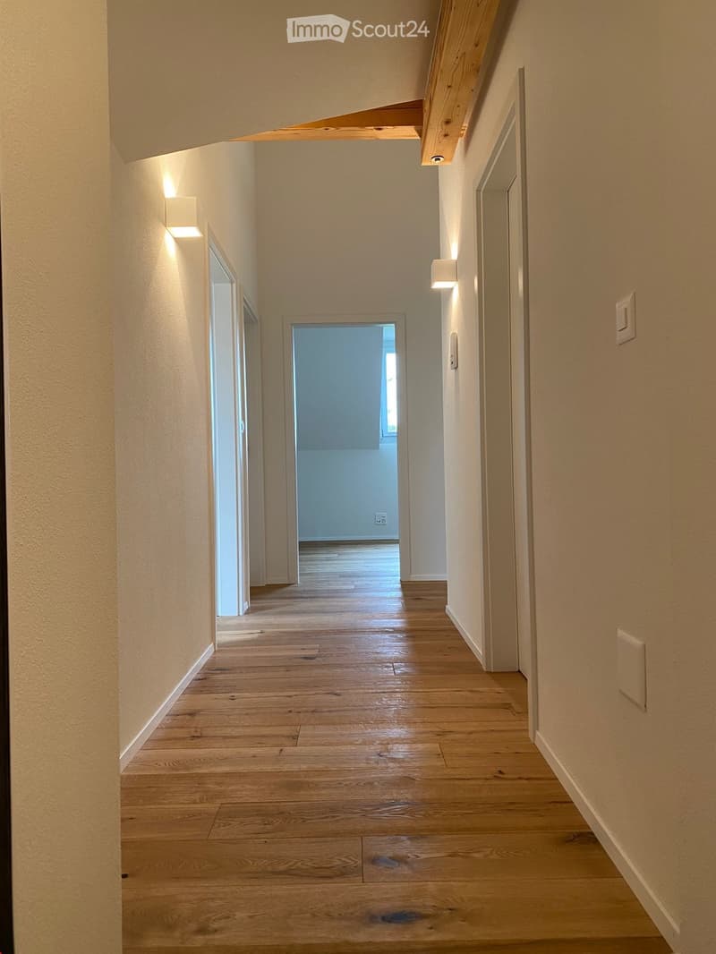 A long, narrow hallway with light wooden flooring and cream-colored walls. The hallway features recessed lighting and a visible doorway at the end leading to another room.