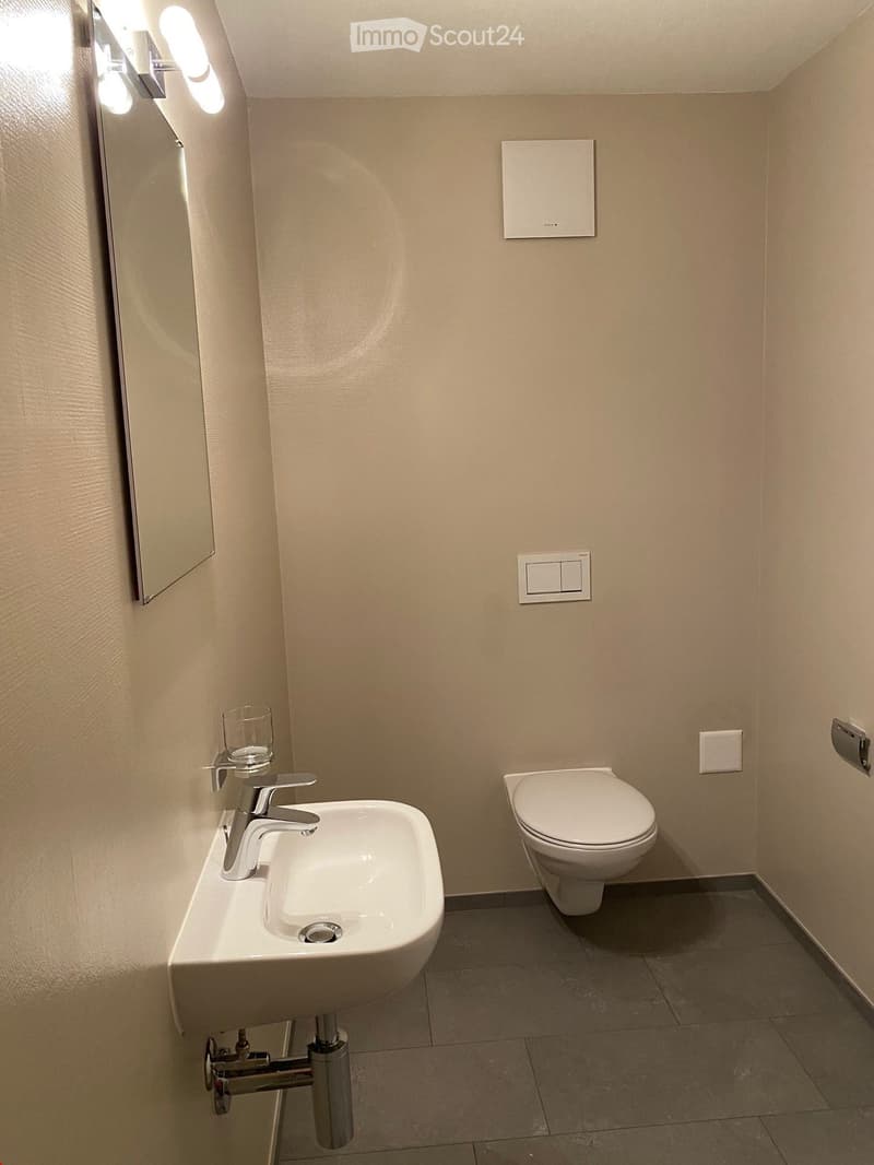 Modern bathroom with a white sink, chrome faucet, toilet, and a wall-mounted mirror with integrated lighting. The walls are light beige, and the floor is tiled in dark gray.
