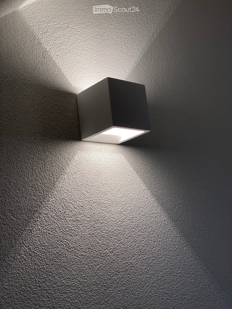 A modern wall-mounted light fixture with a sleek, square design and integrated LED lighting, mounted on a textured gray wall in a hallway.