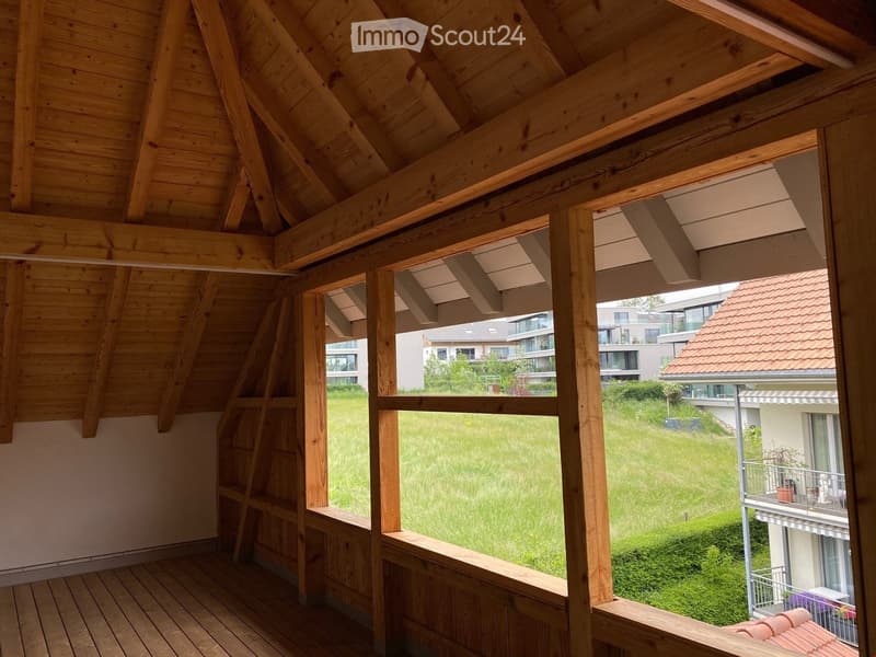 Wooden balcony with slanted wooden ceiling, large windows, and a view of a green lawn and neighboring buildings.