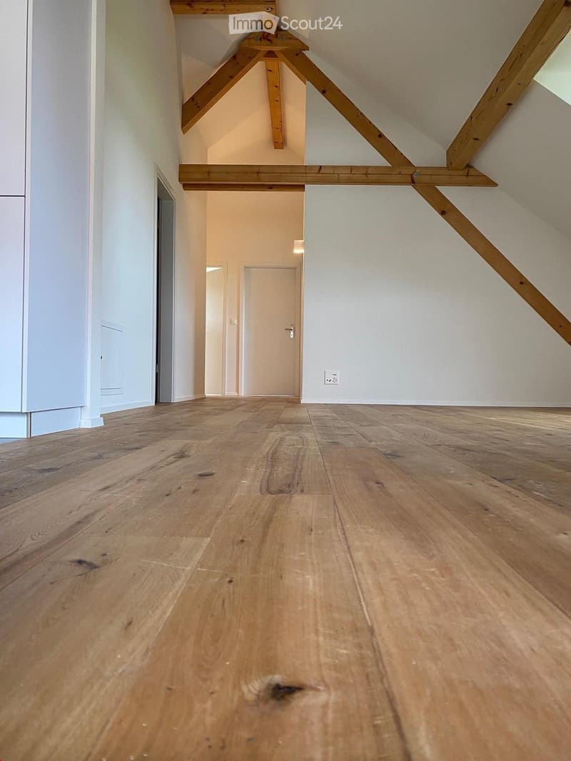 Empty room with wooden flooring, white walls, and exposed wooden beams in the ceiling. There is a closed door at the far end of the room.
