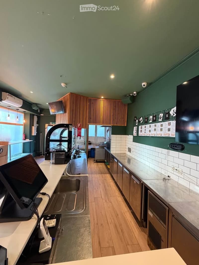 Modern kitchen with wooden flooring, stainless steel appliances, and a large sink area. Countertops are white marble, and there are screens mounted on the walls.