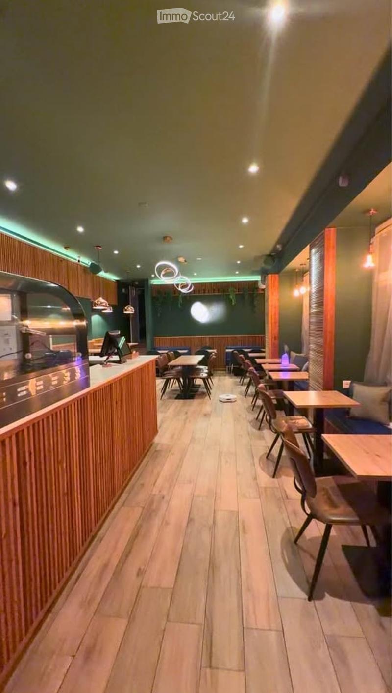 A modern restaurant interior with wooden flooring, a counter on the left, and rows of tables and chairs arranged neatly. The space features warm lighting and a stylish design.