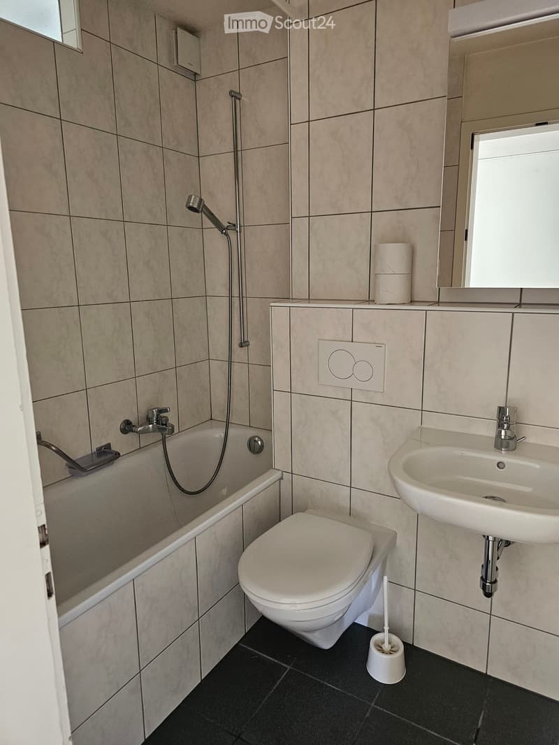 Modern bathroom with beige tiled walls, black tiled floor, bathtub, toilet, sink, and showerhead.