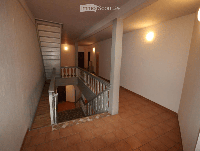 Spacious hallway with a staircase leading to an upper level, wooden railings, warm lighting, and tiled flooring.