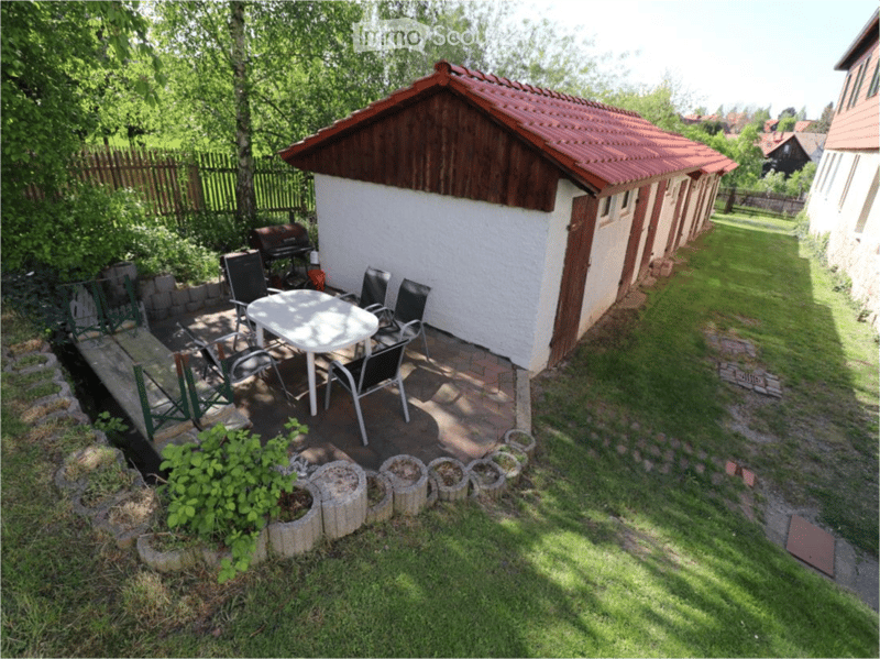 Outdoor patio area with a small table and chairs, surrounded by a wooden fence and greenery. A small wooden shed is visible in the background.