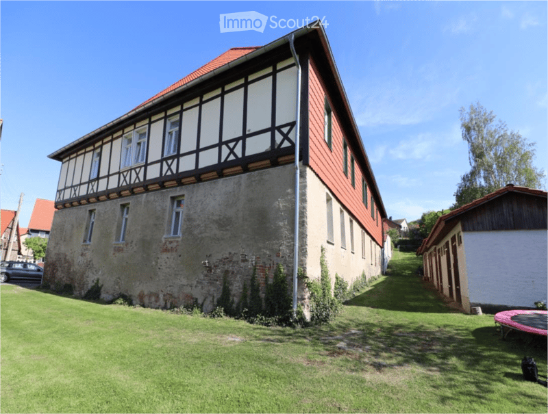 Two-story house with red-tiled roof, white and grey facade, multiple windows, and a small outbuilding. The house features a mix of modern and traditional architectural elements, with a grassy lawn in front.