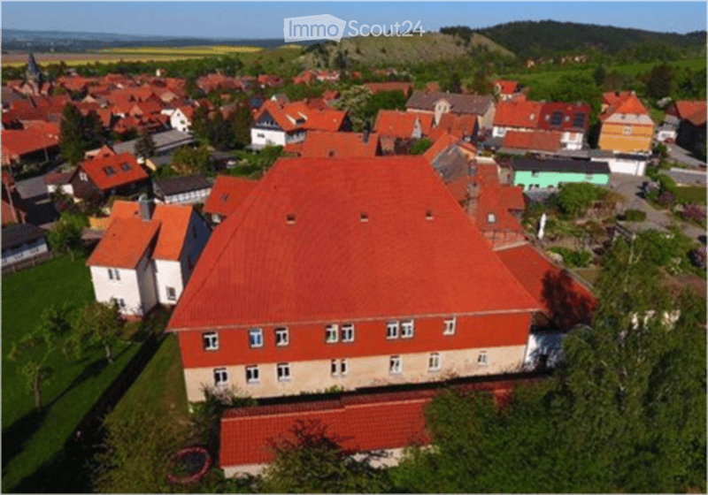 Aerial view of a large, red-roofed building surrounded by smaller houses and green fields in a rural village setting.