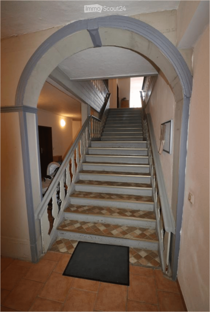 A narrow hallway with a staircase leading upwards, featuring a patterned tile floor and a dark doormat at the base of the stairs.