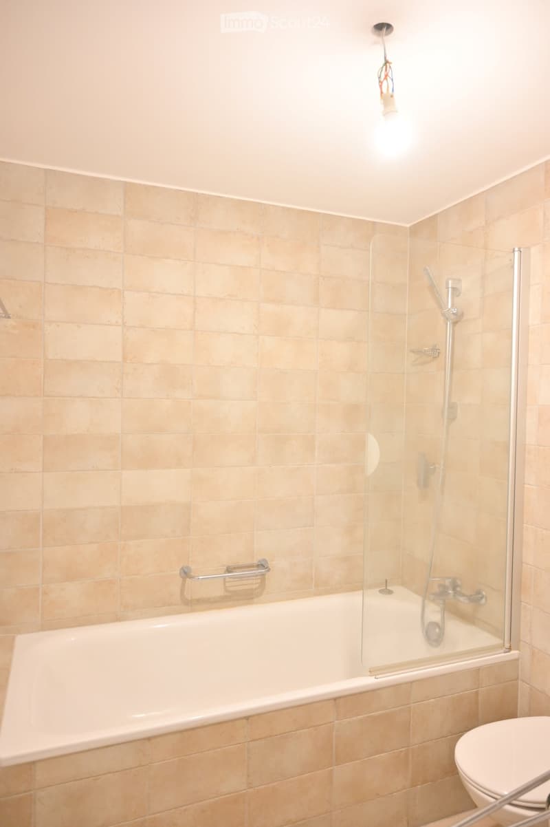 Bathroom with beige tiled walls, a bathtub with a glass shower enclosure, a toilet, and a handrail for support.